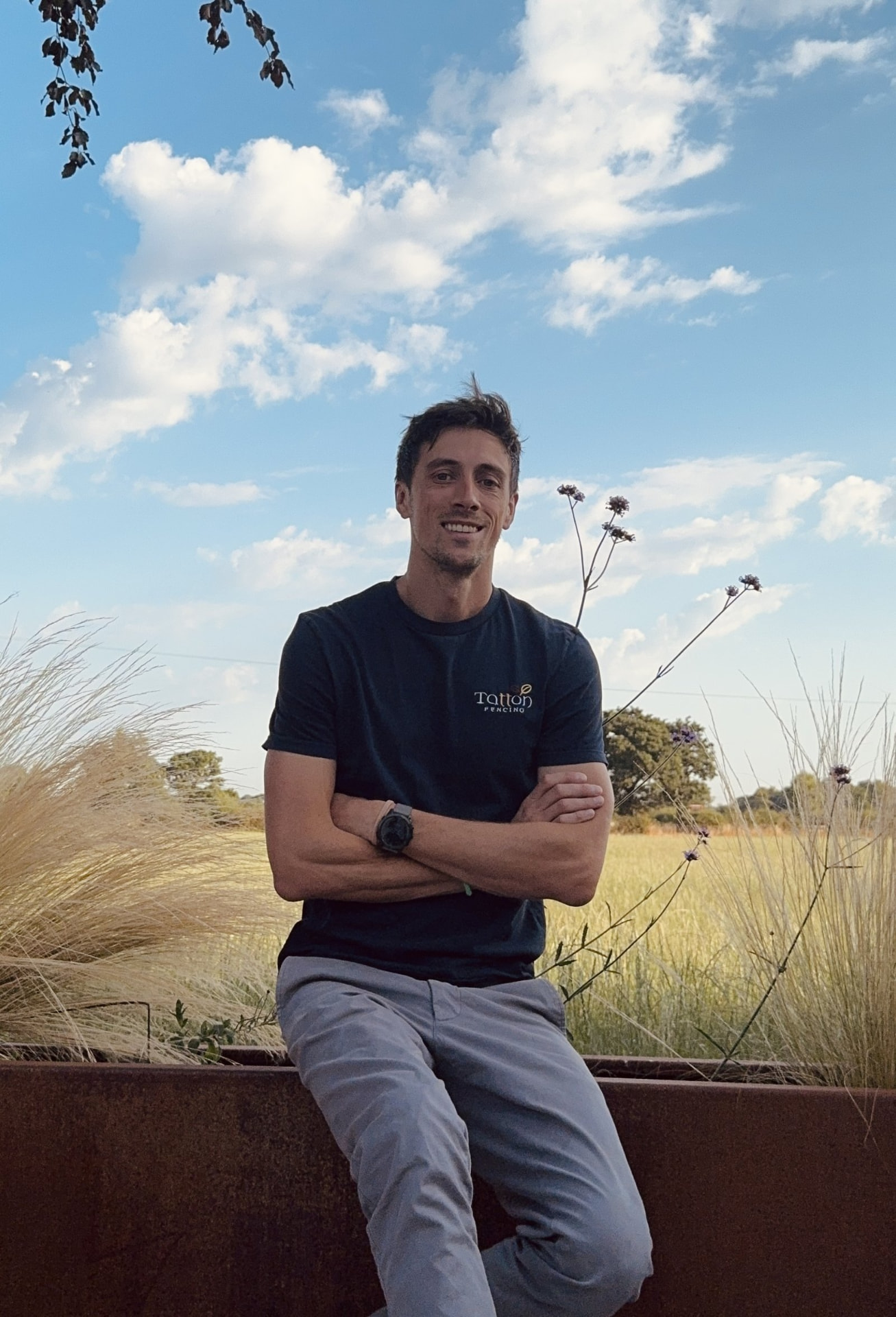 Christopher Vincent, founder of Tatton Fencing, sitting outdoors in a Tatton Fencing t-shirt with Cheshire fields behind him